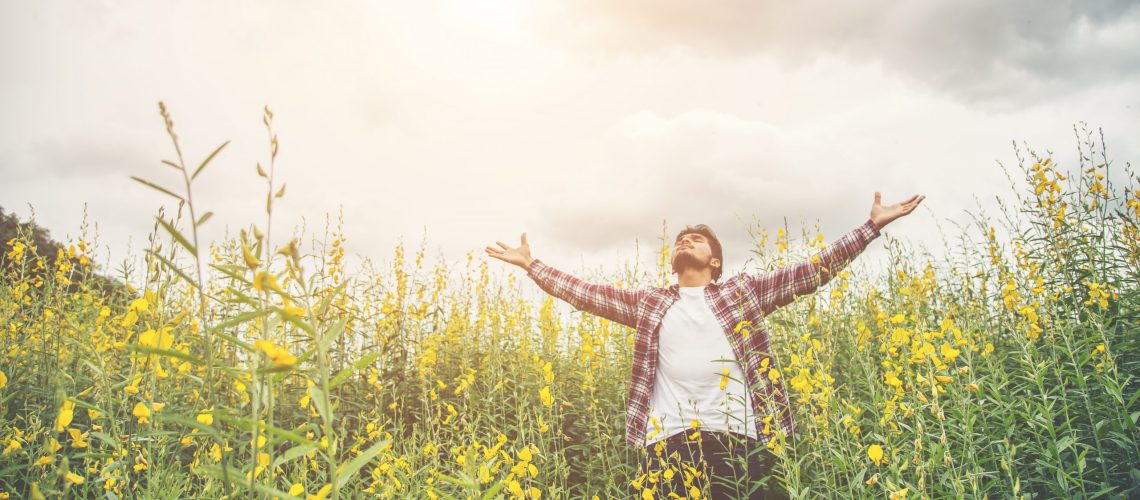 Handsome hipster man standing raising hands in the air in a yellow flower field.